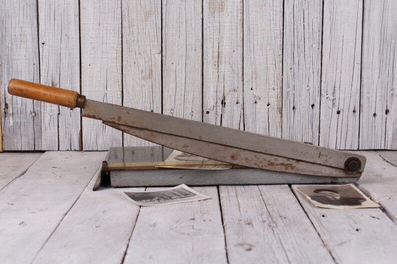 May include: An antique paper cutter with a wooden handle and a metal blade. The cutter is gray and shows signs of wear. Two vintage black and white photographs are on the wooden surface.