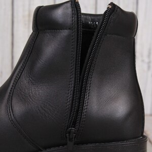 Peut inclure: Gros plan sur une botte en cuir noir avec une fermeture &eacute;clair lat&eacute;rale. La botte a un bout arrondi et un petit talon empil&eacute;. La fermeture &eacute;clair est ouverte, r&eacute;v&eacute;lant l'int&eacute;rieur. Le cuir semble lisse et poli.