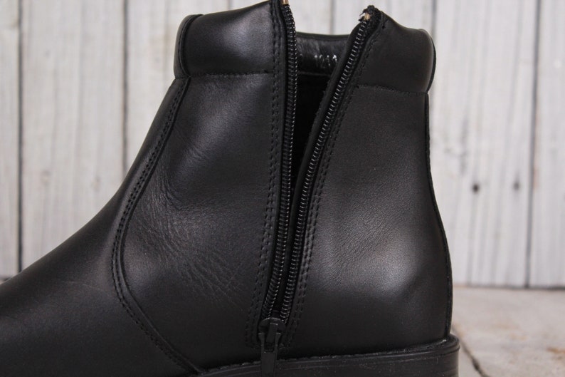 Peut inclure: Gros plan sur une botte en cuir noir avec une fermeture &eacute;clair lat&eacute;rale. La botte a un bout arrondi et un petit talon empil&eacute;. La fermeture &eacute;clair est ouverte, r&eacute;v&eacute;lant l'int&eacute;rieur. Le cuir semble lisse et poli.