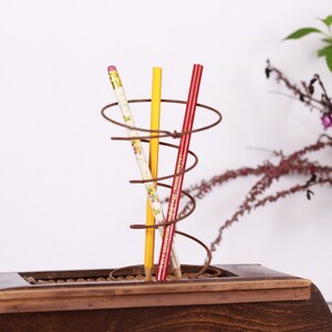 May include: A wooden desk organizer with a spiral wire pencil holder. Three pencils are in the holder: a floral-patterned pencil, a yellow pencil, and a red pencil. The organizer is on a wooden base.