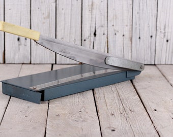 May include: A gray metal paper cutter with a silver blade and a yellow handle. The cutter is sitting on a wooden surface.