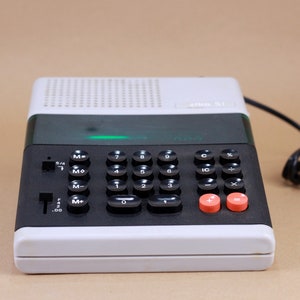 Peut inclure: Une calculatrice blanche et noire avec un écran vert et un bouton rouge et noir. La calculatrice est équipée d'un cordon.