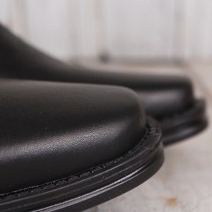 Peut inclure: Gros plan sur deux chaussures en cuir noir. Les chaussures ont une surface lisse et brillante et un bout arrondi. Les coutures autour de la semelle sont visibles. L'arri&egrave;re-plan est une surface en bois floue et de couleur claire.