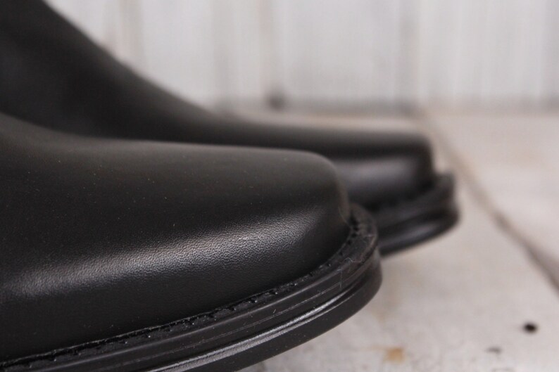 Peut inclure: Gros plan sur deux chaussures en cuir noir. Les chaussures ont une surface lisse et brillante et un bout arrondi. Les coutures autour de la semelle sont visibles. L'arri&egrave;re-plan est une surface en bois floue et de couleur claire.
