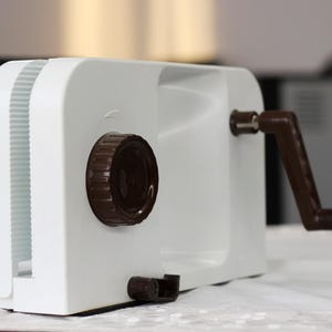 Pode incluir: Máquina de fazer massa de plástico branco com uma manivela e um botão marrons. A máquina de fazer massa tem um lado com nervuras e um lado liso.