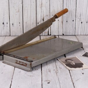 May include: A vintage gray paper cutter with a wooden handle, resting on a white wooden surface. Two black and white photographs are next to the cutter. The blade is angled upwards.
