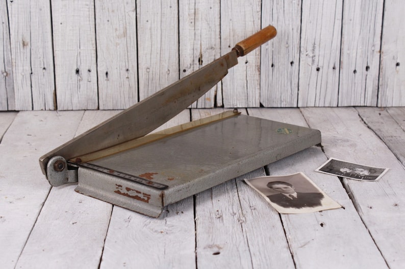 May include: A vintage gray paper cutter with a wooden handle, resting on a white wooden surface. Two black and white photographs are next to the cutter. The blade is angled upwards.