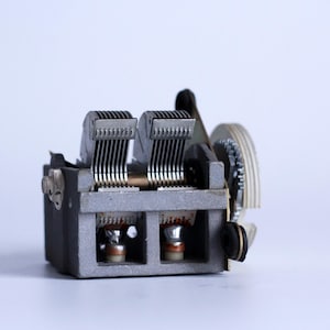 Może przedstawiać: Zabytkowy kondensator zmienny, element stosowany w obwodach radiowych. Urządzenie wykonane jest z metalu i posiada wiele równoległych płytek, pokrętło strojenia oraz różne punkty połączeń. Kolor jest głównie szary z srebrnymi i miedzianymi akcentami.