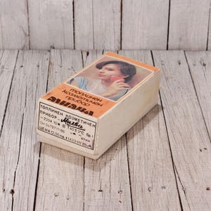 May include: Vintage cosmetic device box with a woman's image on the front. The box is rectangular, with text in a foreign language. The box is beige with orange accents and sits on a weathered wooden surface.