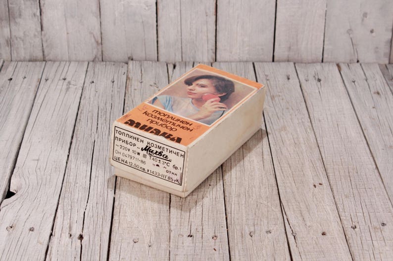May include: Vintage cosmetic device box with a woman's image on the front. The box is rectangular, with text in a foreign language. The box is beige with orange accents and sits on a weathered wooden surface.