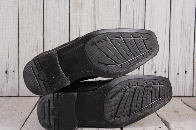 Peut inclure: Deux chaussures de ville en cuir noir avec des semelles textur&eacute;es sont empil&eacute;es sur une surface en bois clair. Les semelles ont un motif g&eacute;om&eacute;trique pour l'adh&eacute;rence. Les chaussures sont probablement neuves ou en excellent &eacute;tat.