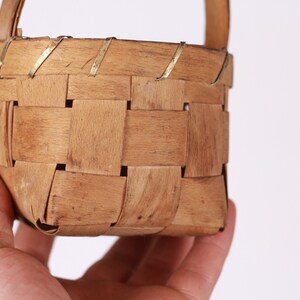 Puede incluir: Una peque&ntilde;a cesta cuadrada tejida con asa. La cesta est&aacute; hecha de tiras de madera marr&oacute;n claro, tejidas en un patr&oacute;n de entramado. Delgadas tiras de metal refuerzan el borde superior. El asa tambi&eacute;n es de madera. La cesta est&aacute; sujeta por una mano.