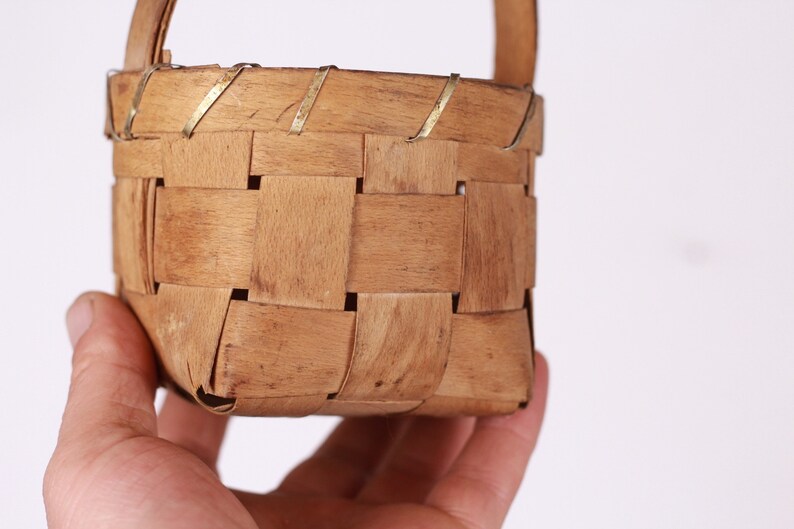 Puede incluir: Una peque&ntilde;a cesta cuadrada tejida con asa. La cesta est&aacute; hecha de tiras de madera marr&oacute;n claro, tejidas en un patr&oacute;n de entramado. Delgadas tiras de metal refuerzan el borde superior. El asa tambi&eacute;n es de madera. La cesta est&aacute; sujeta por una mano.