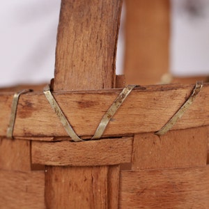 Puede incluir: Primer plano de una peque&ntilde;a cesta de madera tejida con asa. La cesta es de madera marr&oacute;n claro y tiene forma rectangular. Delgadas tiras de metal refuerzan los bordes y las fijaciones del asa.