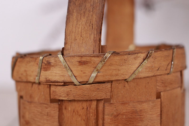 Puede incluir: Primer plano de una peque&ntilde;a cesta de madera tejida con asa. La cesta es de madera marr&oacute;n claro y tiene forma rectangular. Delgadas tiras de metal refuerzan los bordes y las fijaciones del asa.