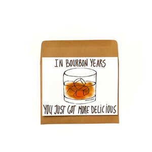 K&ouml;nnte beinhalten: Eine wei&szlig;e Karte mit braunem Papierumschlag, die eine handgezeichnete Illustration eines Glases Whisky mit Eisw&uuml;rfeln zeigt. Der Text auf der Karte lautet "In bourbon years you just got more delicious."