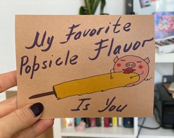 May include: A brown card with the text "My favorite popsicle flavor is you" written in purple ink. A cartoon pig is lying on top of a yellow popsicle.
