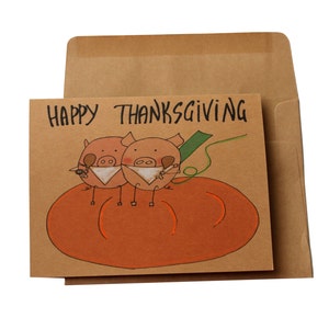 May include: A brown card with the text "Happy Thanksgiving" written in black. Two cartoon pigs are sitting on a large orange pumpkin. The pigs are wearing white bibs and holding a drumstick in their mouths.