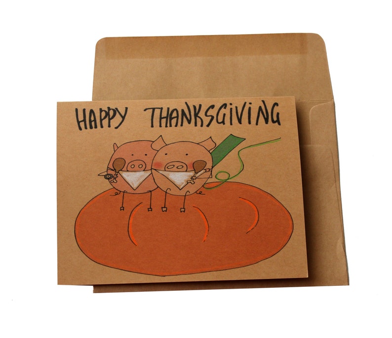 May include: A brown card with the text "Happy Thanksgiving" written in black. Two cartoon pigs are sitting on a large orange pumpkin. The pigs are wearing white bibs and holding a drumstick in their mouths.