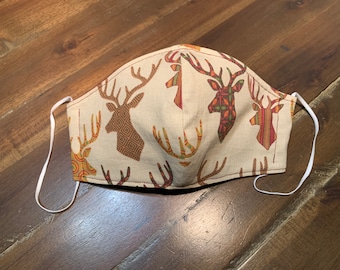May include: A beige face mask with a repeating pattern of brown, orange, and green deer heads. The mask has white elastic ear loops.