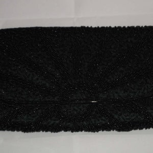Può includere: Una pochette nera con perline di forma rettangolare. La pochette presenta una chiusura a patta con una piccola chiusura color argento. La superficie è ricoperta da piccole perline scure disposte in un motivo strutturato.