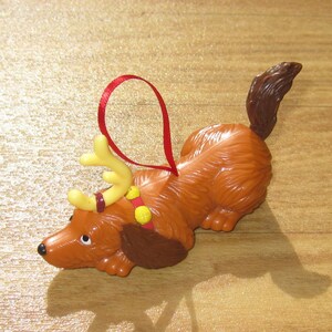 May include: A brown dog figurine ornament with a long tail, wearing yellow plastic reindeer antlers and a red collar with yellow bells. A thin red ribbon loop is attached to the top of the dog for hanging as a holiday decoration.