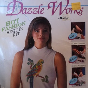 Peut inclure: Un haut sans manches blanc avec un motif de perroquet à paillettes. L'emballage indique "Dazzle Works HOT FASHION SEQUIN KIT" et comprend des instructions pour le transfert thermocollant, la couture et le collage de paillettes.