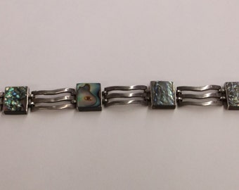 May include: A silver bracelet featuring square abalone shell inlays. The bracelet has a series of square abalone shell pieces connected by three wavy silver links. The abalone shells display iridescent colors of blue, green, and purple.