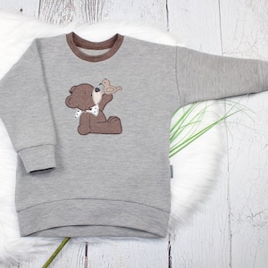 Könnte beinhalten: Grauer Pullover mit einem braunen Bären- und Vogel-Applikation. Der Bär hält einen kleinen Vogel in seiner Pfote. Der Pullover hat einen braunen gerippten Kragen.