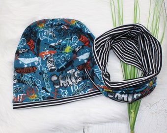 May include: A blue beanie with a black and white stripe trim. The beanie has a graffiti-style print with the word "SKATE" and other words and images. A black and white striped neck warmer with the same print is also shown.