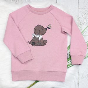 Könnte beinhalten: Ein altrosa Langarm-Sweatshirt mit einer Grafik eines braunen Welpen, der ein weißes Bandana mit schwarzen Punkten trägt und einen kleinen Schmetterling betrachtet. Das Kleidungsstück hat einen Rundhalsausschnitt, gerippte Bündchen und ein kleines Markenetikett.