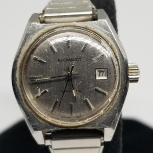 May include: A vintage Movado automatic wristwatch with a silver-tone case and band. The watch face is gray with silver hands and markers, and a date window at the 3 o'clock position. The watch is marked "Swiss Made".