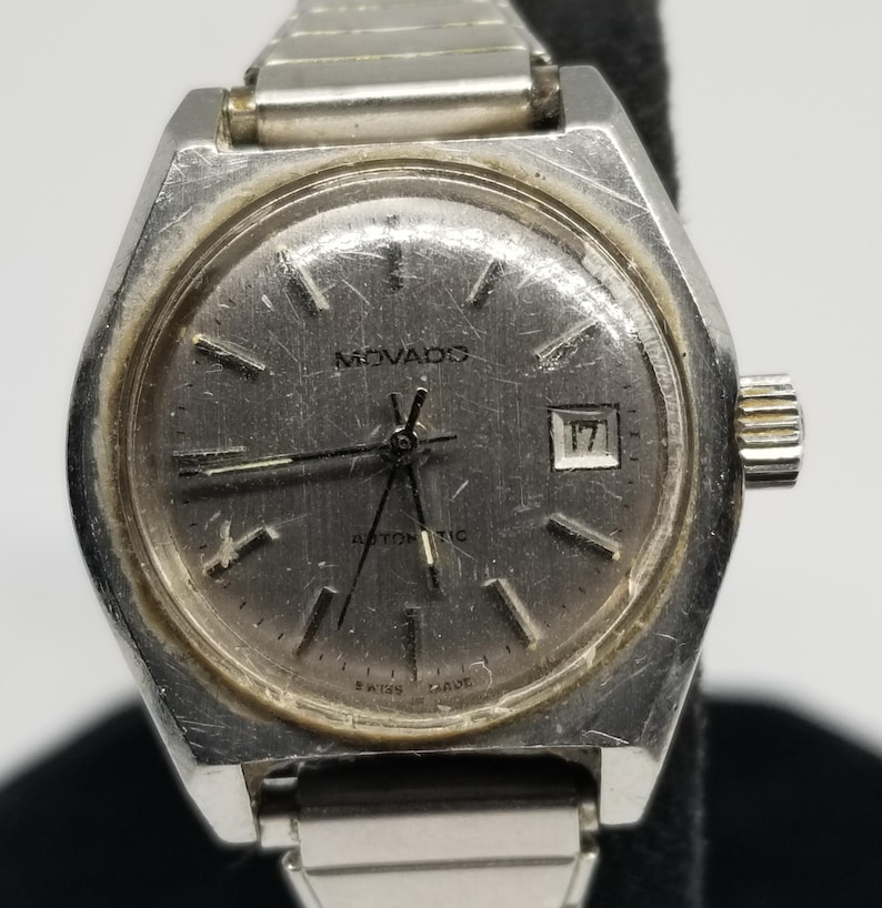 May include: A vintage Movado automatic wristwatch with a silver-tone case and band. The watch face is gray with silver hands and markers, and a date window at the 3 o'clock position. The watch is marked "Swiss Made".