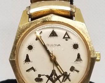 Puede incluir: Un reloj Bulova vintage con caja y correa doradas. La esfera del reloj es de color crema con símbolos negros y la marca Bulova. El reloj presenta una caja hexagonal y una correa texturizada.
