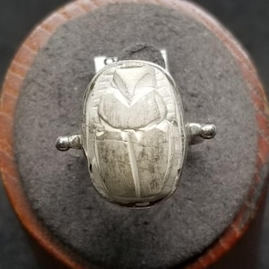 May include: A silver ring featuring a detailed scarab beetle design. The ring has a rounded, oval-shaped face with intricate carvings depicting the beetle's features. The band extends to small, rounded accents on either side of the beetle.