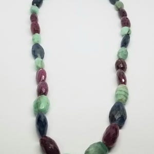 May include: A necklace featuring a variety of polished gemstones in shades of green, blue, and burgundy. The stones are strung together with a gold clasp. The necklace is displayed against a white background.