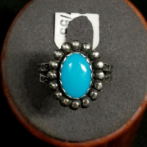 May include: A silver ring with a bright turquoise oval stone at the center. The ring has a beaded border around the stone and a double-banded design. The ring is displayed on a gray surface.