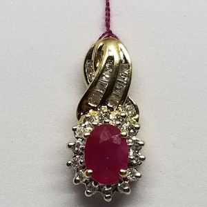 May include: A gold pendant featuring a large oval ruby gemstone surrounded by small diamonds. The pendant has a twisted gold design with additional diamond accents. A maroon string is attached for hanging.