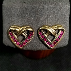 May include: A pair of gold-toned heart-shaped earrings. Each earring features a heart design with a row of small, round, pink gemstones. The earrings are displayed on a gray surface.