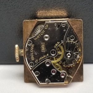 Puede incluir: Un movimiento de reloj vintage Bulova Watch Co., que muestra intrincados engranajes y componentes. El marco dorado rodea una esfera negra con el texto "Bulova Watch Co. U.S.A. 17 Jewels". Los detalles mec&aacute;nicos del reloj son visibles, destacando su artesan&iacute;a.