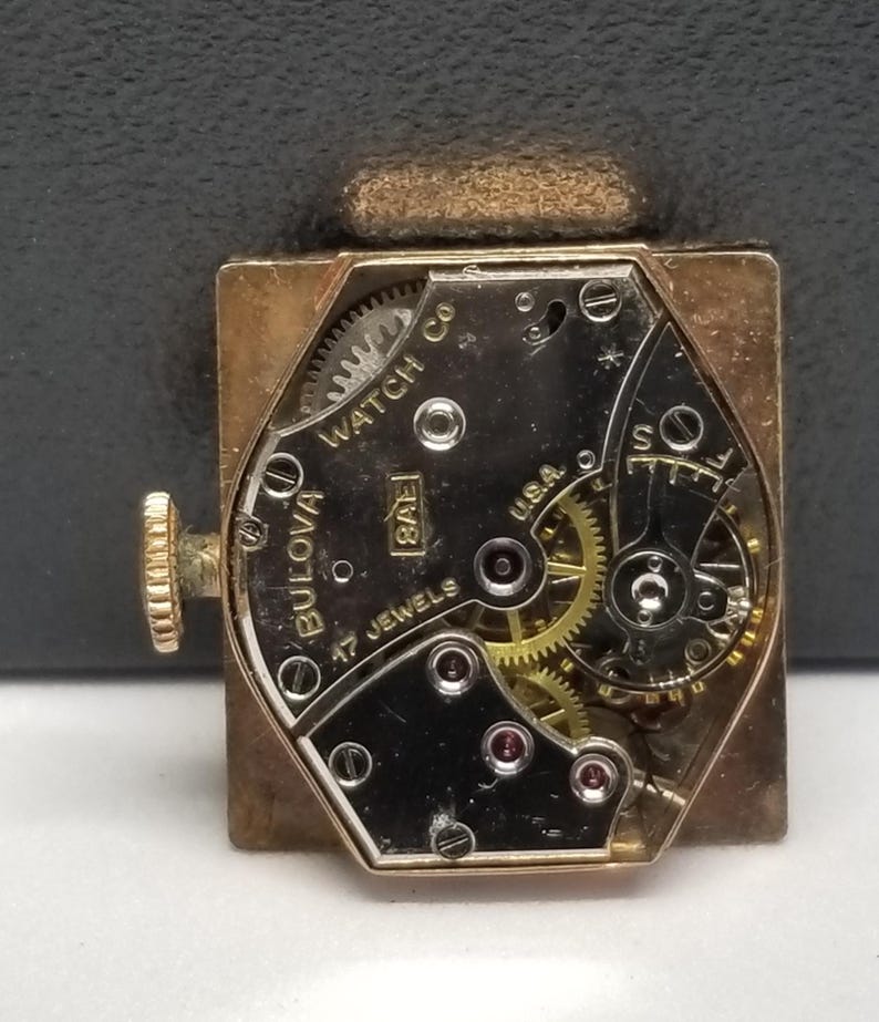 Puede incluir: Un movimiento de reloj vintage Bulova Watch Co., que muestra intrincados engranajes y componentes. El marco dorado rodea una esfera negra con el texto "Bulova Watch Co. U.S.A. 17 Jewels". Los detalles mec&aacute;nicos del reloj son visibles, destacando su artesan&iacute;a.