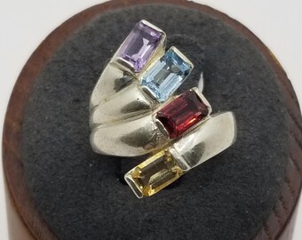 May include: A silver ring with four colorful gemstones. The ring has a twisted design and features a purple, blue, red, and yellow gemstone.