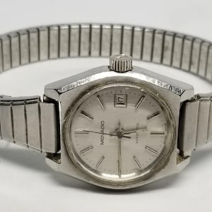 May include: A silver-toned Movado automatic wristwatch with a rectangular face and a date display. The watch has a silver band and a textured crown. The watch face has the Movado logo and the word "automatic".