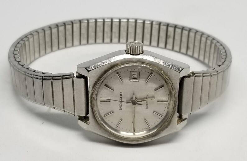 May include: A silver-toned Movado automatic wristwatch with a rectangular face and a date display. The watch has a silver band and a textured crown. The watch face has the Movado logo and the word "automatic".