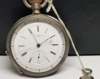 Puede incluir: Un reloj de bolsillo plateado con esfera blanca, números romanos negros y un pequeño dial de segundos. El reloj tiene una corona dorada y una llave sujeta con una cuerda. La esfera del reloj dice "John Forrest & Co. Liverpool".