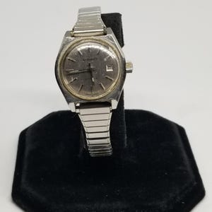 May include: A vintage silver-toned watch with a rectangular face and a silver band. The watch face has a light-colored background with black hands and hour markers. The watch is displayed on a black velvet stand.