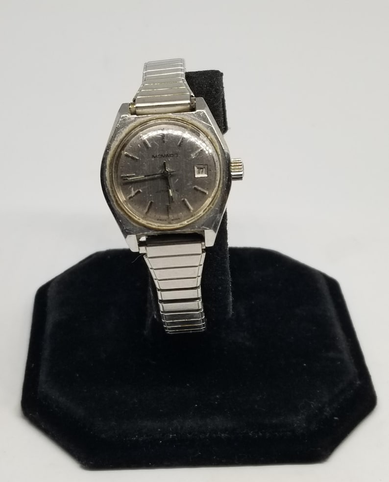 May include: A vintage silver-toned watch with a rectangular face and a silver band. The watch face has a light-colored background with black hands and hour markers. The watch is displayed on a black velvet stand.