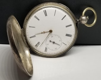 Puede incluir: Un reloj de bolsillo antiguo abierto, con esfera blanca, números romanos negros y un pequeño dial de segundos. La caja plateada está parcialmente abierta, revelando un diseño grabado. El reloj tiene una tija de cuerda y un lazo para una cadena.