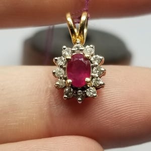 May include: A gold pendant with a central oval ruby surrounded by a halo of small diamonds. The pendant is suspended from a thin, purple string, and is held between two fingers.