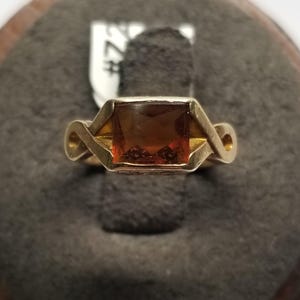 May include: A gold ring featuring a square-cut, amber-colored gemstone set in a geometric frame. The band displays a stylized, infinity-like design on both sides of the central stone. A small white tag with the text #724 is visible behind the ring.